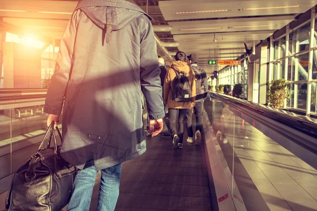 Një udhëtar ecën nëpër një korridor aeroporti të ndriçuar nga dielli me valixhe, drejt pushimeve.