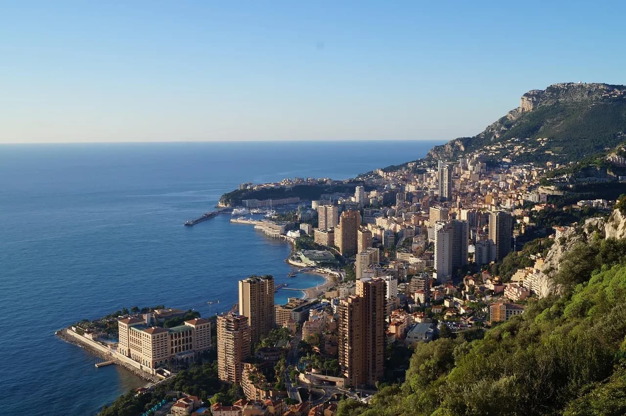 Aerial view of Monte Carlo's towers and coastline on the Mediterranean, with steep green hills behind.