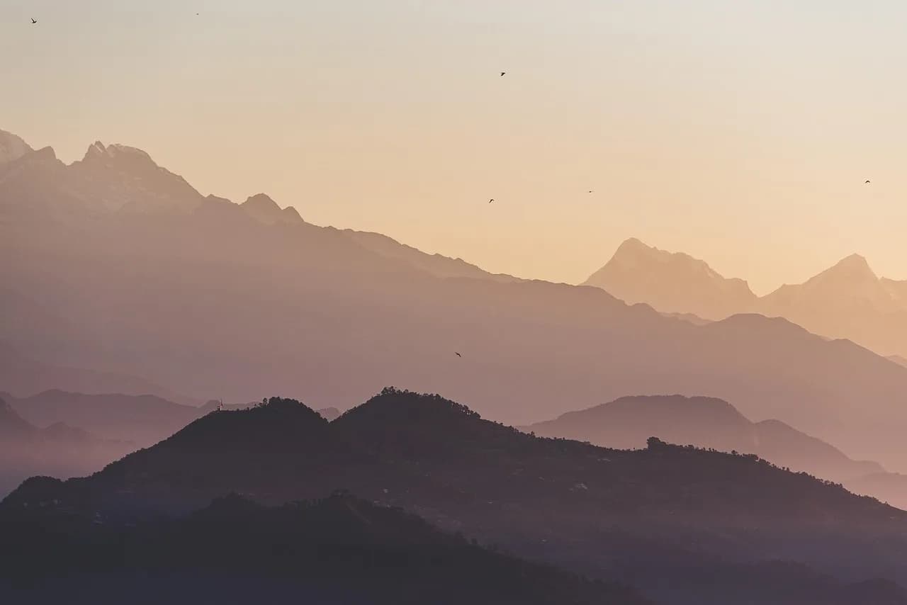 Siluetat e shtresuara të maleve shkëlqejnë ngrohtësisht në perëndim të diellit, duke ftuar për udhëtim.
