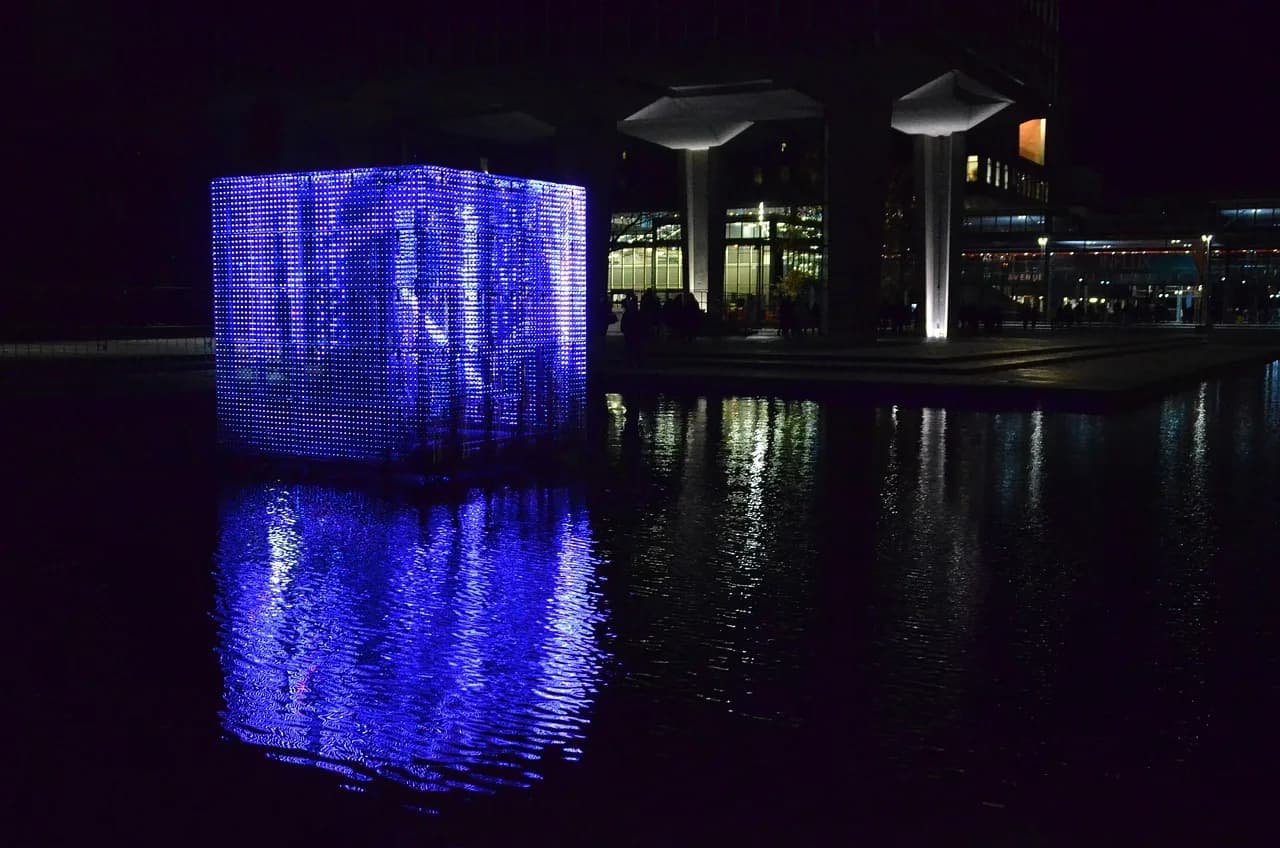 A glowing blue light-art cube reflected in the water at night, Eindhoven.