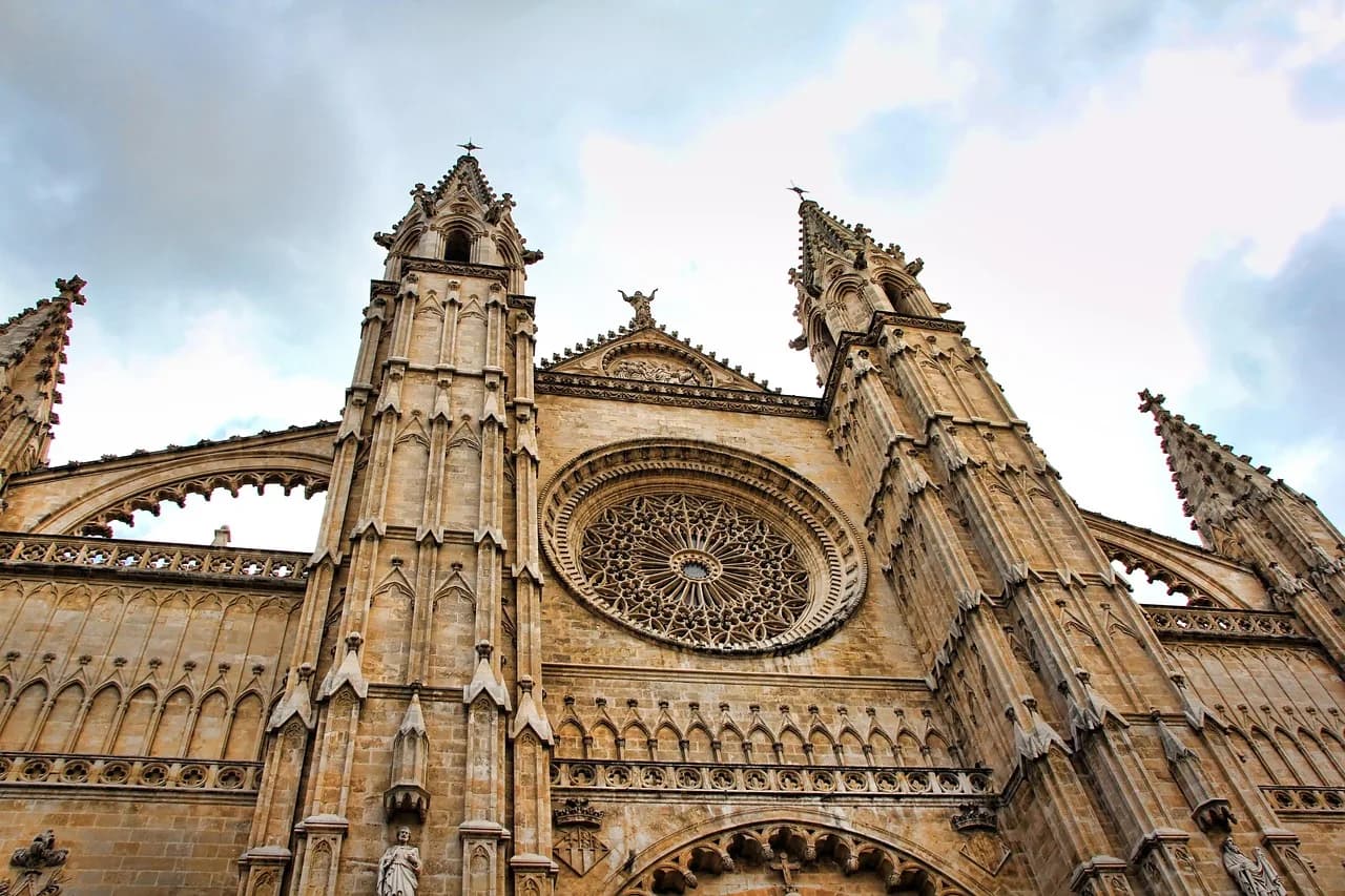 Gothic facade and rose window of Palma Cathedral seen from below against a cloudy sky.