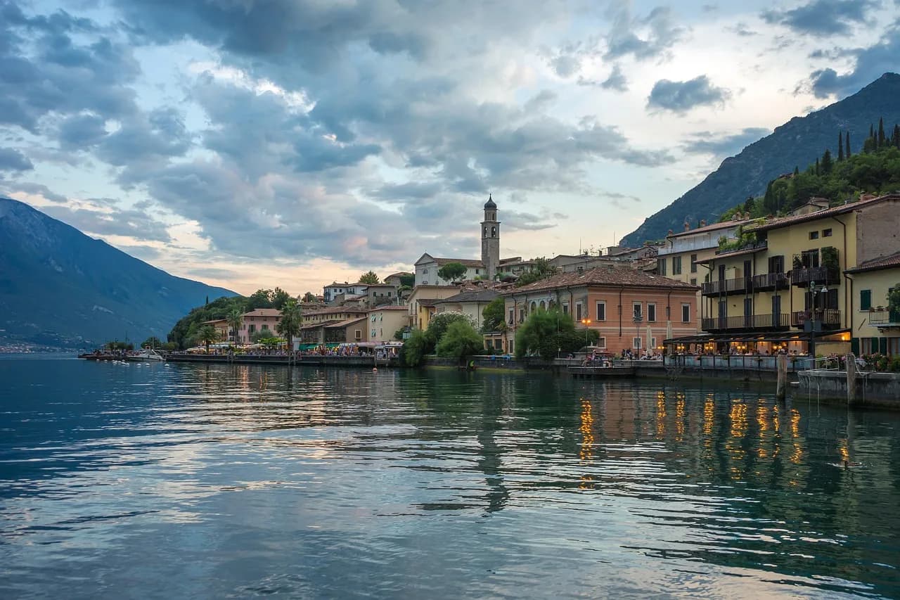 Un borgo portuale sul Lago di Garda con campanile e montagne riflesse nell'acqua calma.