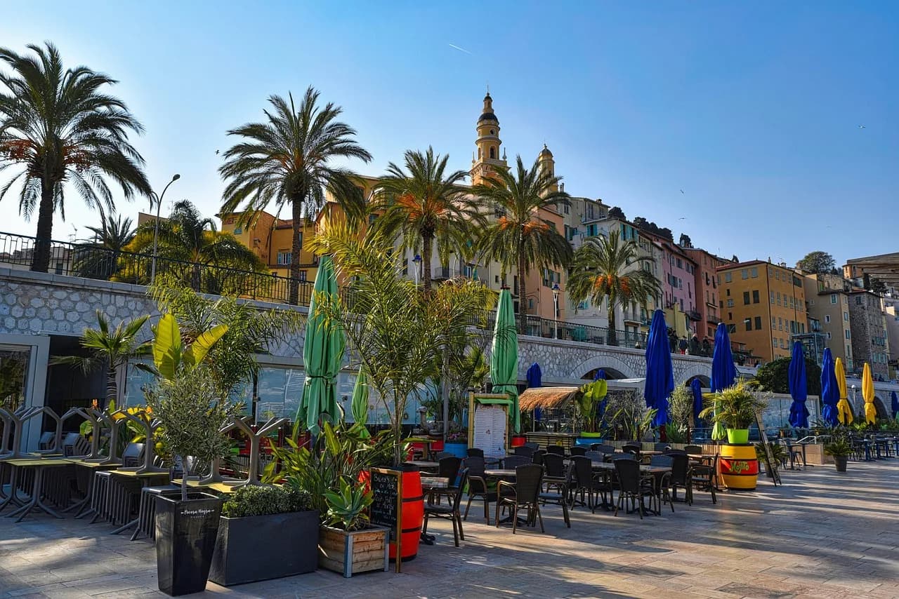 A sunny restaurant terrace in Menton under palm trees, with pastel buildings and a bell tower above.