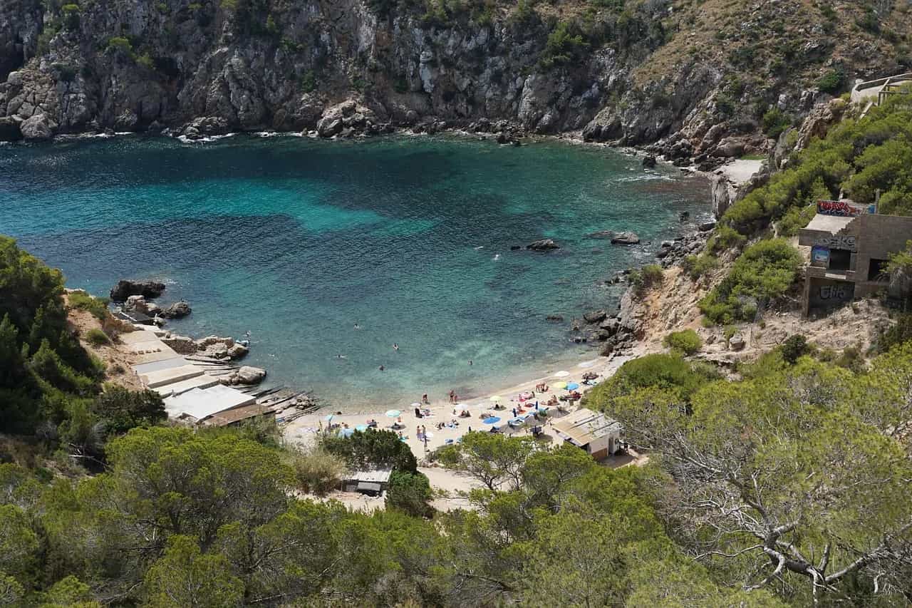A turquoise cove with a small pebble beach below pine-covered cliffs on Ibiza.
