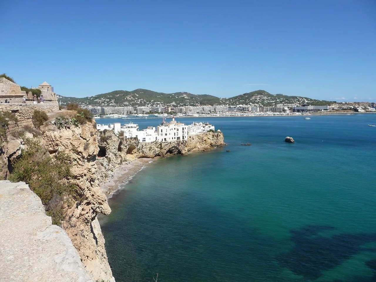 White buildings at the water's edge beside a deep blue Mediterranean bay on Ibiza.